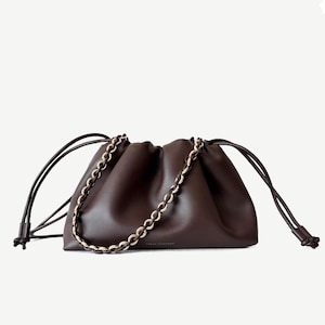 Könnte beinhalten: Eine dunkelbraune Ledertasche in Beutelform mit geraubtem Verschluss und langen Kordelzügen. Die Tasche verfügt über einen dekorativen, goldfarbenen Kettenriemen, der über die Vorderseite fällt, sowie ein kleines goldenes Logo unten in der Mitte.