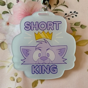 Może przedstawiać: Jasnoniebieska odznaka z fioletowym napisem "SHORT KING", przedstawiająca kreskówkowego kota w złotej koronie. Odznaka ma metalowy klips na górze.