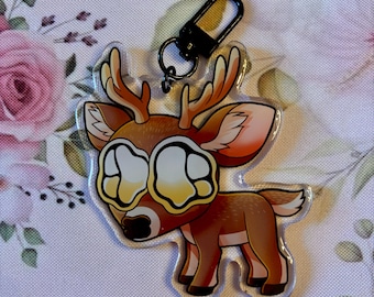 May include: A clear acrylic keychain featuring a cartoon deer with large, expressive eyes. The deer has brown fur, antlers, and a black nose. The keychain includes a black clasp for attaching to keys or bags. The background is a floral pattern.