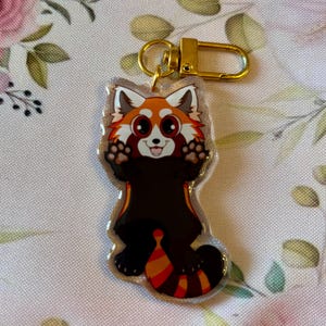 May include: A clear acrylic keychain featuring a cartoon red panda with a gold-colored clasp. The red panda has a white face, orange fur, and a striped tail. The keychain is on a floral background.