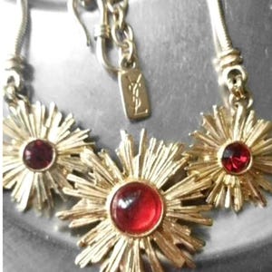 Op de afbeelding: Een goudkleurige ketting met drie zonnestraalvormige hangers, elk in het midden voorzien van een ronde, dieprode cabochonsteen. De ketting bevat een metalen label met het YSL-logo en een haaksluiting bevestigd aan een getextureerde ketting.
