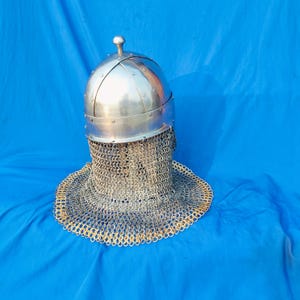 May include: A silver helmet with a chainmail coif. The helmet has a rounded top and a pointed tip. The chainmail coif is made of small metal rings linked together.