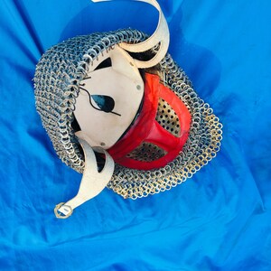May include: A medieval-style helmet with a chainmail coif and a red leather face mask. The helmet is made of white leather and has a small eye hole. The helmet is lying on a blue surface.