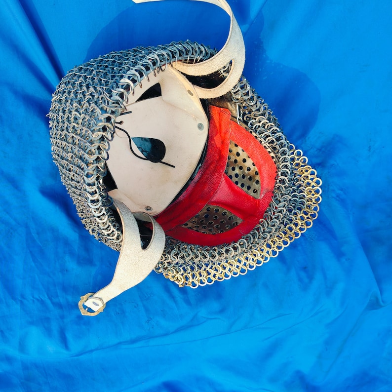 May include: A medieval-style helmet with a chainmail coif and a red leather face mask. The helmet is made of white leather and has a small eye hole. The helmet is lying on a blue surface.
