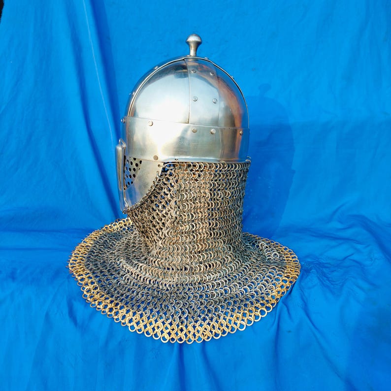 May include: A silver helmet with a chainmail coif. The helmet has a pointed top and a visor with a small hole in it. The chainmail coif is made of small metal rings.