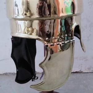 Puede incluir: Una réplica dorada de un casco romano con una protección de mejilla con bisagras. El casco está en un soporte de madera.