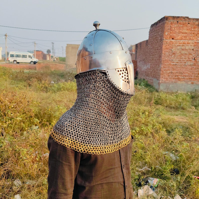 May include: A silver helmet with a chainmail coif. The helmet has a pointed top and a face mask with a small opening for the eyes. The coif is made of small metal rings linked together.