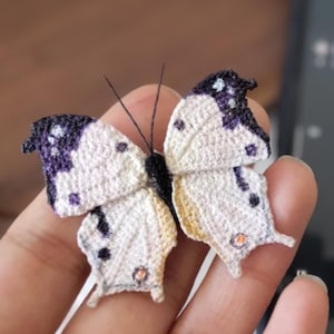 May include: A handcrafted butterfly with white and purple wings, detailed with black accents and small orange embellishments. The butterfly is made of a textured material, possibly fabric or yarn, and has delicate antennae.