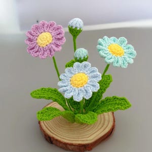 Pode incluir: Um arranjo de flores de crochê feito à mão com margaridas em tons de roxo, azul e água-marinha com centros amarelos. As flores são colocadas em caules verdes com folhas verdes e montadas em uma fatia de madeira.