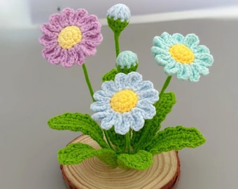 May include: A handmade crochet flower arrangement featuring daisies in shades of purple, blue, and aqua with yellow centers. The flowers are set on green stems with green leaves, and are mounted on a wooden slice.