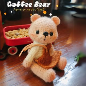 Pode incluir: Um urso de peluche de crochê vestindo uma roupa marrom e branca. O urso tem um nariz rosa e olhos pretos. O texto "Coffee Bear have a nice day :)" é visível no fundo.