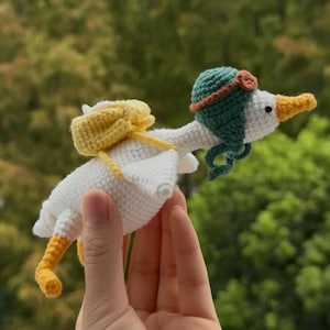 May include: A handmade white crochet goose with yellow feet and beak, wearing a green aviator hat with brown goggles and a yellow backpack. The whimsical design is perfect for a nursery or child's room.