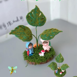 May include: A whimsical miniature scene featuring a crocheted green leaf canopy over two small figurines. One figurine wears a blue hooded coat and holds a red umbrella. The other wears a pink hooded coat. The scene includes small red and white mushrooms.