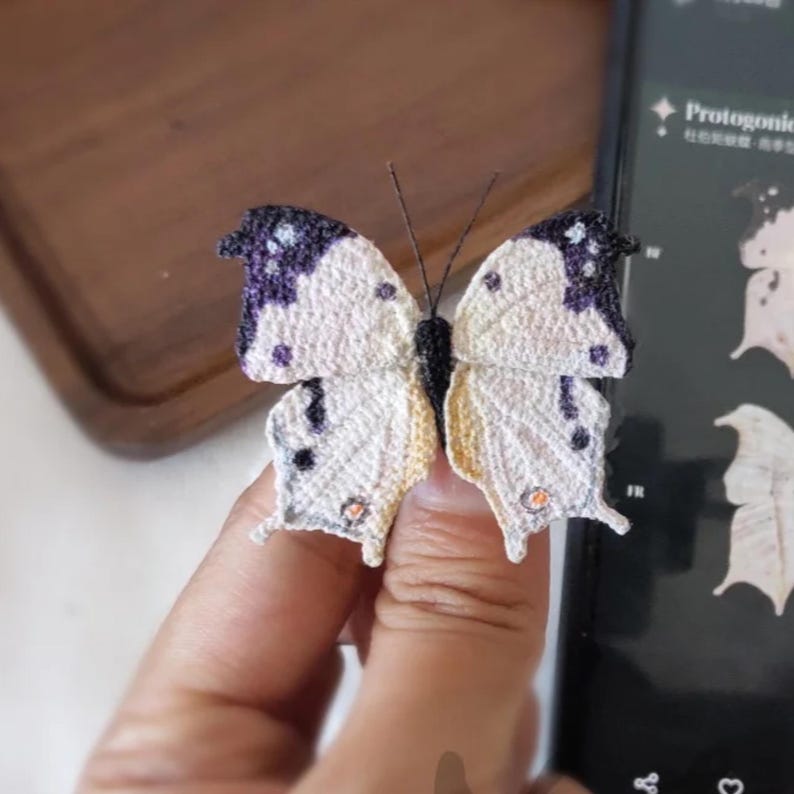 May include: A handcrafted butterfly, featuring a detailed crochet design. The butterfly has white wings with purple and black accents, and a yellow gradient near the body. The antennae are thin and black. The butterfly is held by a hand.