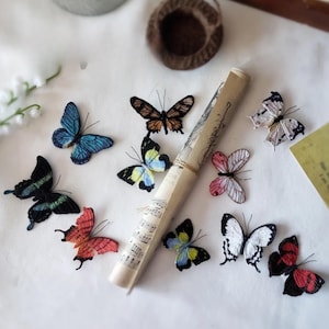 May include: A collection of embroidered butterfly decorations in various colors, including blue, black, red, and white. A rolled scroll with musical notes is also present. The butterflies are arranged on a white surface.