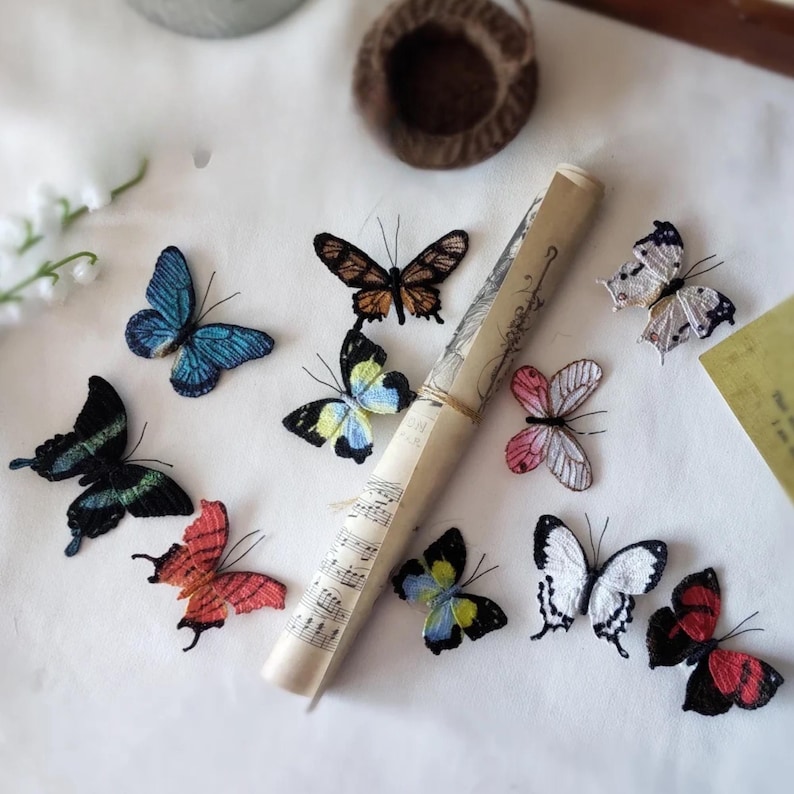 May include: A collection of embroidered butterfly decorations in various colors, including blue, black, red, and white. A rolled scroll with musical notes is also present. The butterflies are arranged on a white surface.