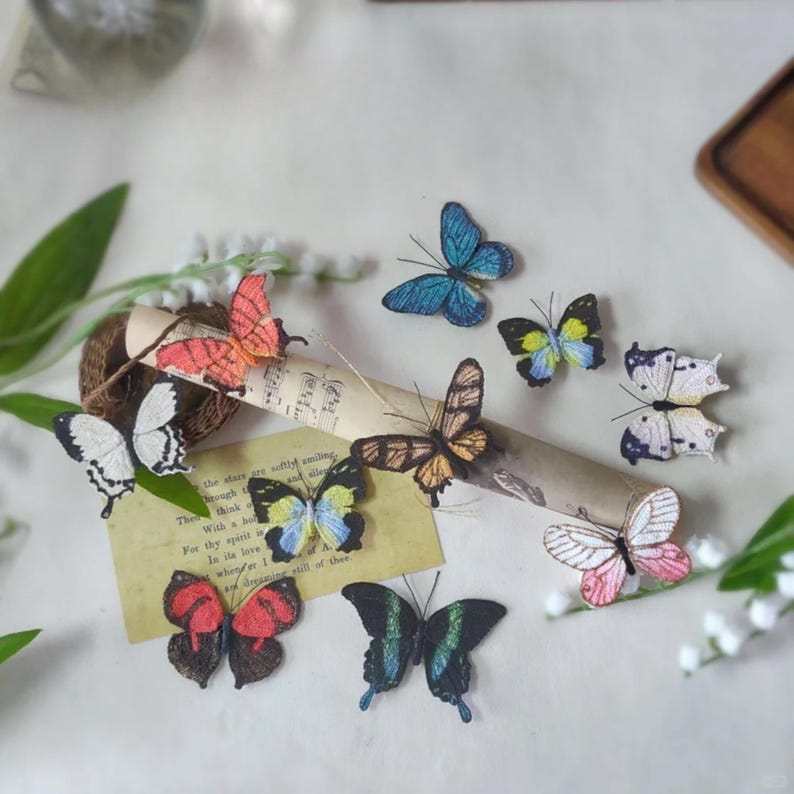 May include: A collection of colorful butterfly embellishments in various sizes and designs. The butterflies feature shades of blue, pink, orange, black, and white, with intricate details. Some butterflies are resting on a vintage paper with text.