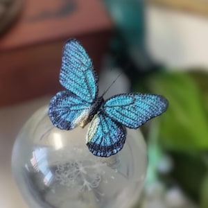 May include: A handcrafted crochet butterfly with blue and black wings, resting on a clear glass sphere. The butterfly's wings display intricate patterns and textures, showcasing the detailed craftsmanship. The background is softly blurred, highlighting the butterfly.