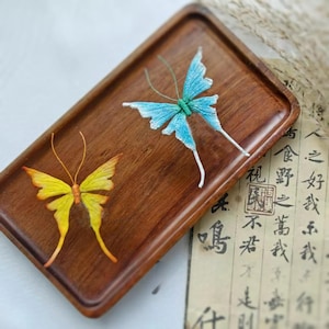May include: Two handcrafted butterfly ornaments. One is yellow and orange, the other is blue and white. They are displayed on a wooden tray with Chinese calligraphy.