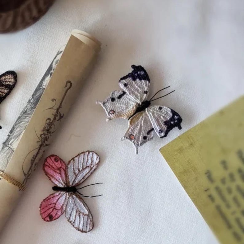 May include: Embroidered butterfly decorations in white, pink, and black, with delicate wing patterns. A rolled parchment and a yellowed paper are also in the image. The butterflies appear to be handmade, possibly for crafts or decor.