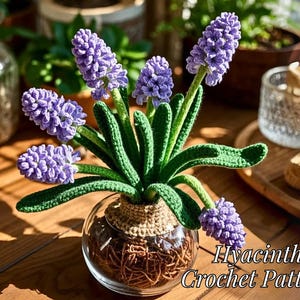 Puede incluir: Un arreglo de flores de jacinto de ganchillo en un jarrón de vidrio. Las jacintas son moradas y las hojas son verdes. El jarrón está lleno de material marrón que parece tierra. El texto "Hyacinth Crochet Pattern" está escrito debajo del jarrón.