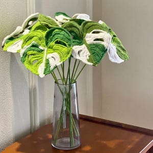 May include: A bouquet of crocheted green and white monstera leaves in a clear glass vase.