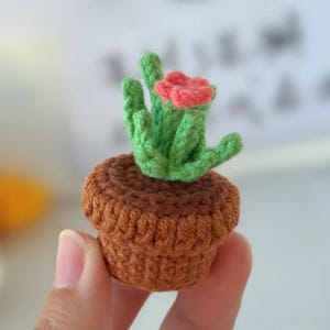 Może przedstawiać: Mały, ręcznie robiony szydełkowy kaktus w brązowej doniczce. Kaktus ma zielone łodygi i czerwony kwiat. Doniczka jest w ciepłym brązowym kolorze, a całość wykonana jest z przędzy.