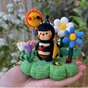 May include: A crocheted bee with a yellow and black striped body, red cheeks, and a black and red ladybug in its hand. The bee is standing on a green crocheted cloud with a yellow sun, a white daisy, and a blue flower.