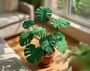 May include: A handcrafted, green crochet Monstera plant in a brown terracotta pot. The plant features large, intricately crocheted leaves with characteristic fenestrations. The pot sits on a wooden surface, with natural light illuminating the scene.
