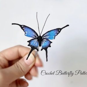 Può includere: Una farfalla all'uncinetto con ali blu e nere. La farfalla presenta dettagli delicati e lunghe antenne. Il testo "Crochet Butterfly Pattern" è scritto in corsivo sotto.