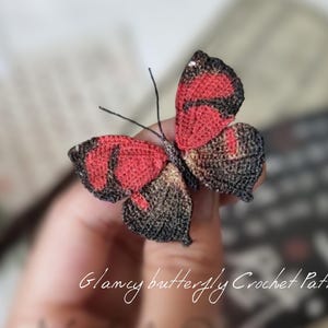 Könnte beinhalten: Ein handgehäkelter Schmetterling mit roten und schwarzen Flügeln und einem Hauch von Gold. Der Schmetterling hat zarte Fühler. Der Text "Glamcy butterfly Crochet Pattern" ist kursiv geschrieben.