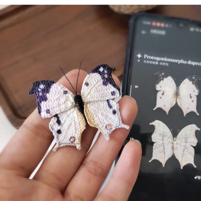 May include: A handcrafted butterfly with white and purple wings, detailed with intricate stitching. The butterfly has delicate antennae and is held in a hand. A phone screen displays a butterfly image.