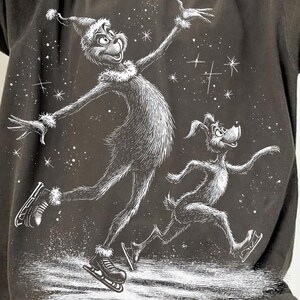 Puede incluir: Camiseta gris oscuro de gran tamaño con una ilustración en blanco y negro de un personaje de dibujos animados y un perro patinando sobre hielo. El diseño gráfico incluye copos de nieve y estrellas, creando un tema invernal.