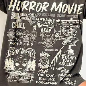 May include: A charcoal grey oversized t-shirt featuring a distressed cream-coloured graphic print. The design includes the text Horror Movie, various horror film references, character illustrations, and iconic movie locations in a collage layout.