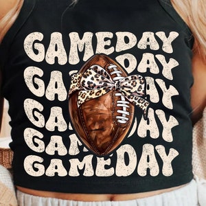 May include: Black sleeveless crop top with the text "GAMEDAY" repeated in a retro font. A brown football with a leopard print bow is centred on the shirt.