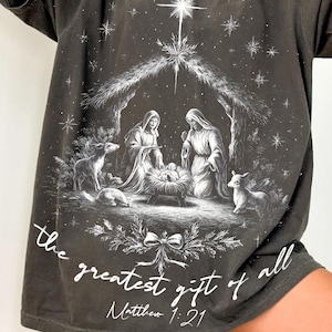 May include: Dark gray t-shirt featuring a Nativity scene with the text "the greatest gift of all" and "Matthew 1:21". The graphic is in white and depicts Mary, Joseph, and baby Jesus in a manger, with stars and animals.