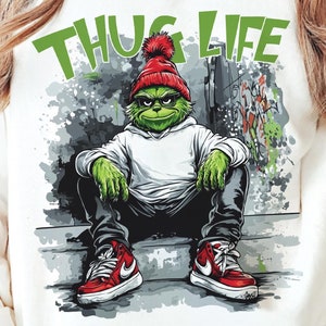 Puede incluir: Sudadera blanca con un gráfico de un personaje verde con gorro rojo y zapatillas rojas, con las palabras "THUG LIFE" en verde sobre el personaje. El personaje está sentado en unos escalones frente a una pared con grafitis.