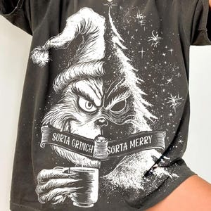 May include: Dark gray t-shirt featuring a white graphic of a Grinch-like character wearing a Santa hat and holding a mug. The text on the banner reads "Sorta Grinch Sorta Merry". The design includes a Christmas tree and stars.