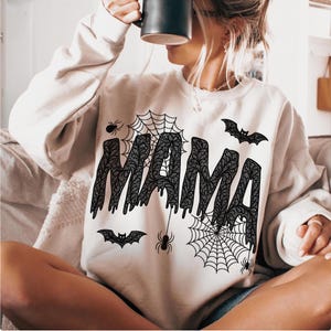May include: A cream-colored sweatshirt with the word "MAMA" in a black, lace-like font. The design includes spiderwebs, spiders, and bats, creating a Halloween theme. The sweatshirt is a casual, long-sleeved garment.