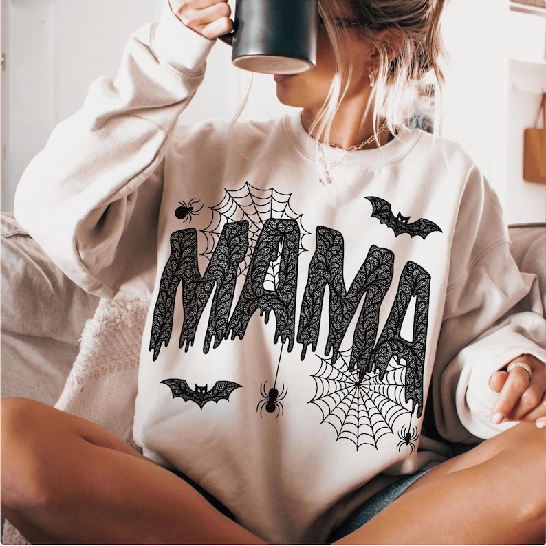 May include: A cream-colored sweatshirt with the word "MAMA" in a black, lace-like font. The design includes spiderwebs, spiders, and bats, creating a Halloween theme. The sweatshirt is a casual, long-sleeved garment.