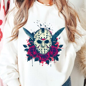 Peut inclure: Sweat-shirt blanc avec un motif graphique. Le motif repr&eacute;sente un masque de hockey, deux couteaux et des roses rouges. Le masque est de couleur cr&egrave;me avec des accents noirs et des d&eacute;tails rouges. Les roses sont d'un rouge profond.