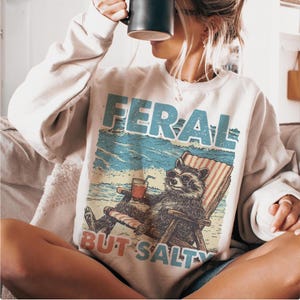 May include: A white sweatshirt featuring a graphic of a raccoon wearing sunglasses and relaxing in a beach chair with a drink. The text "FERAL BUT SALTY" is printed below the raccoon.