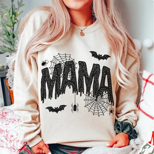 May include: Beige sweatshirt with the word "MAMA" in black, dripping, lace-like letters. The design includes spiderwebs, spiders, and bats, creating a Halloween theme. A cozy and festive garment.