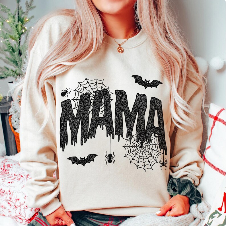 May include: Beige sweatshirt with the word "MAMA" in black, dripping, lace-like letters. The design includes spiderwebs, spiders, and bats, creating a Halloween theme. A cozy and festive garment.