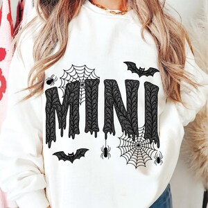May include: White sweatshirt with the word "MINI" in a black, dripping font, decorated with spiderwebs, spiders, and bats. The design has a Halloween theme.