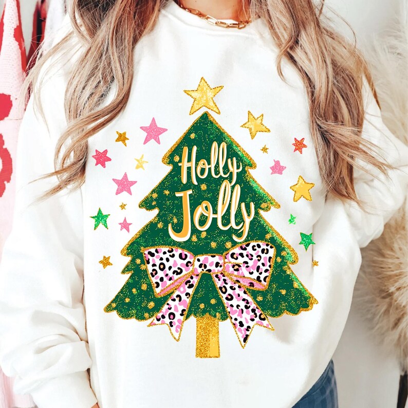 May include: White sweatshirt featuring a green Christmas tree design with the words "Holly Jolly" in white. The tree has a gold star on top and a pink and black leopard print bow. Colorful stars surround the tree.