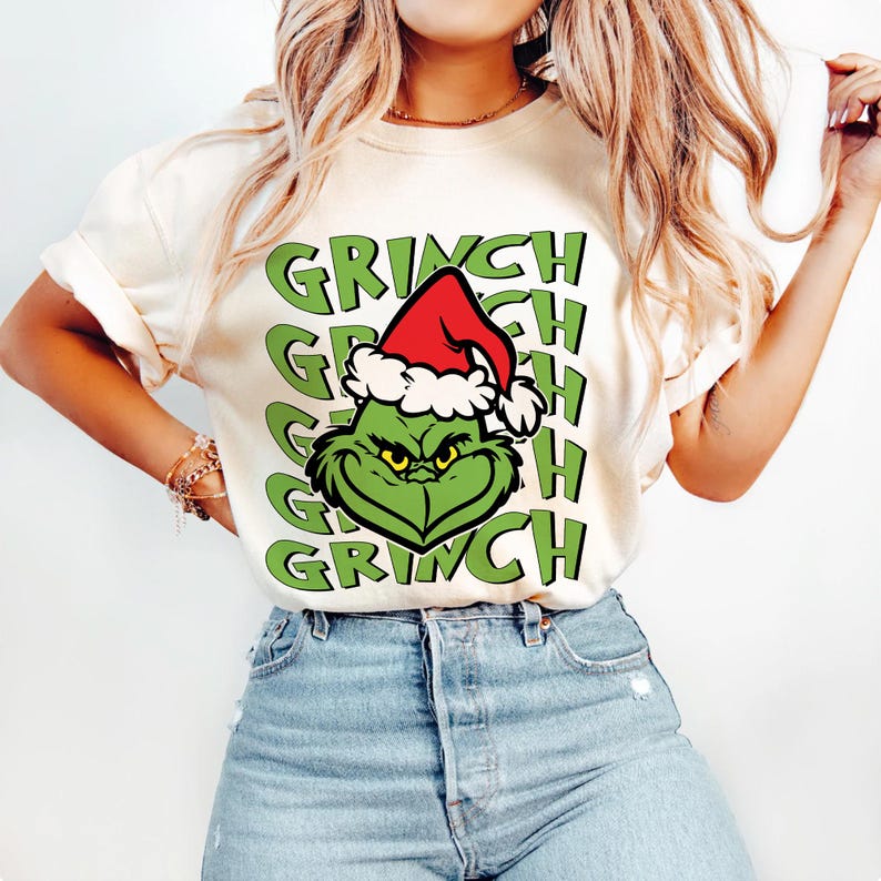 Puede incluir: Camiseta color crema con la cara verde del Grinch con un gorro de Pap&aacute; Noel rojo con ribete blanco. La palabra "GRINCH" est&aacute; apilada verticalmente en letras verdes. La camiseta es de estilo casual.