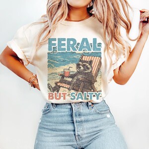 May include: A white t-shirt featuring a vintage-style graphic of a raccoon wearing sunglasses and relaxing in a beach chair with a drink. The text "FERAL BUT SALTY" is printed beneath the raccoon.