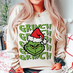 Puede incluir: Sudadera beige con la cara verde del Grinch con un gorro rojo de Pap&aacute; Noel. La palabra "GRINCH" est&aacute; impresa en verde varias veces. Una sudadera con tem&aacute;tica navide&ntilde;a.