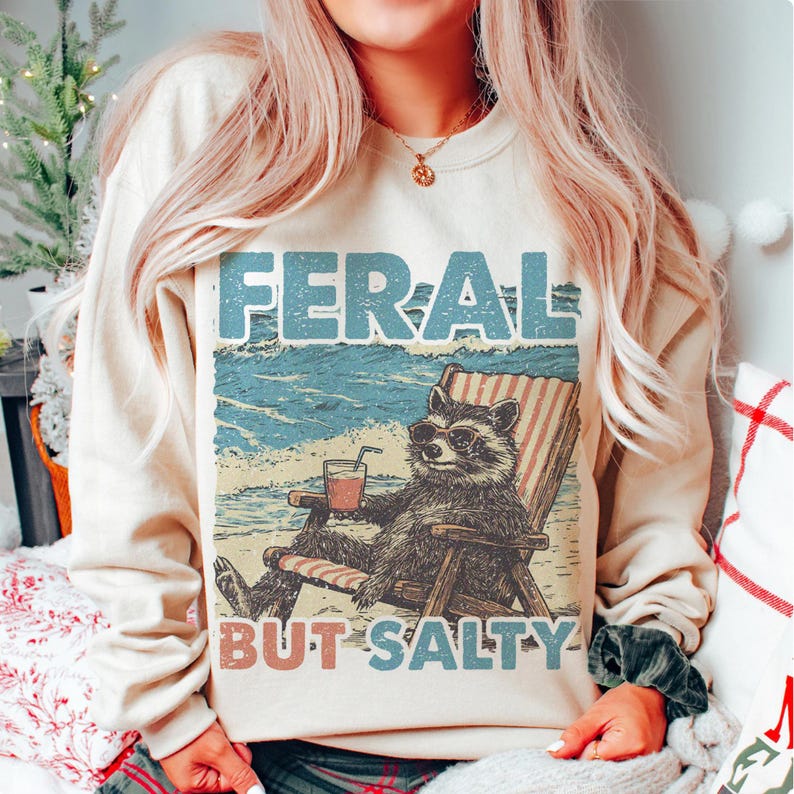 May include: A cream-coloured sweatshirt featuring a graphic of a raccoon wearing sunglasses and relaxing in a beach chair with a drink. The text "FERAL BUT SALTY" is printed below the raccoon.